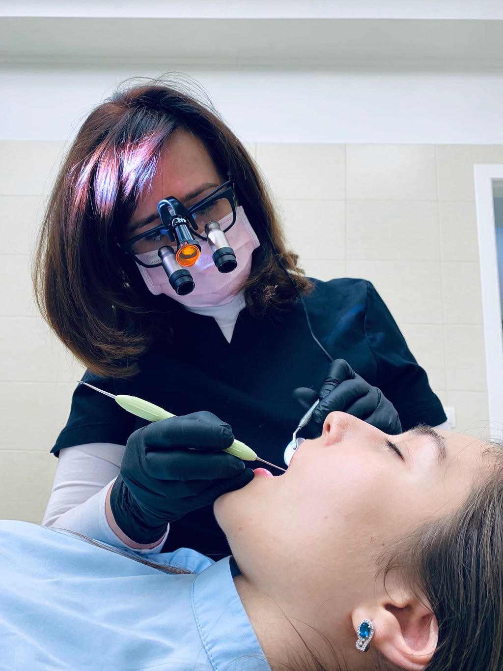 A dental technician with serious eyewear uses metal tools in a patient’s mouth.