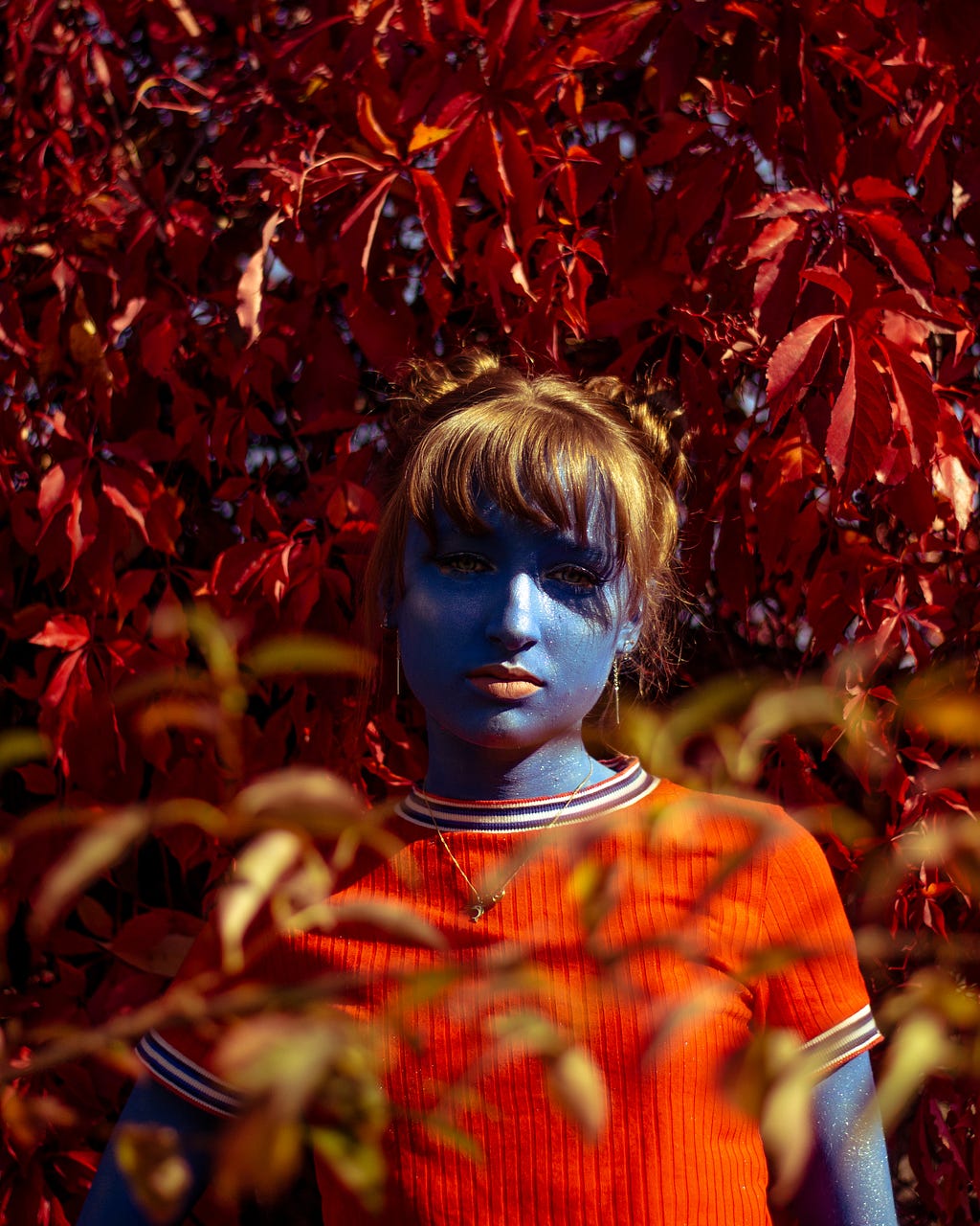 A photo portrait of a woman with blue skin and strawberry orange hair with red leaves surrounding her.