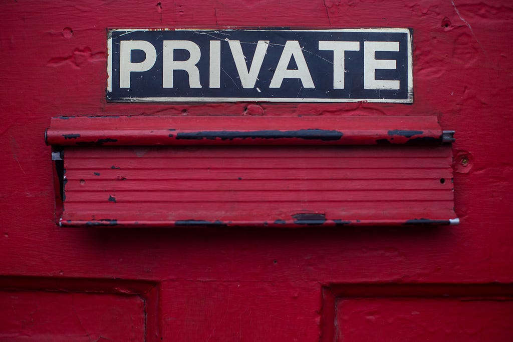 This is a photo of a red door with a black and white “PRIVATE” sign and a red mail slot. The door is painted red and has a black and white “PRIVATE” sign on it. The mail slot is red and appears to be old and weathered. The background is a red wall.
