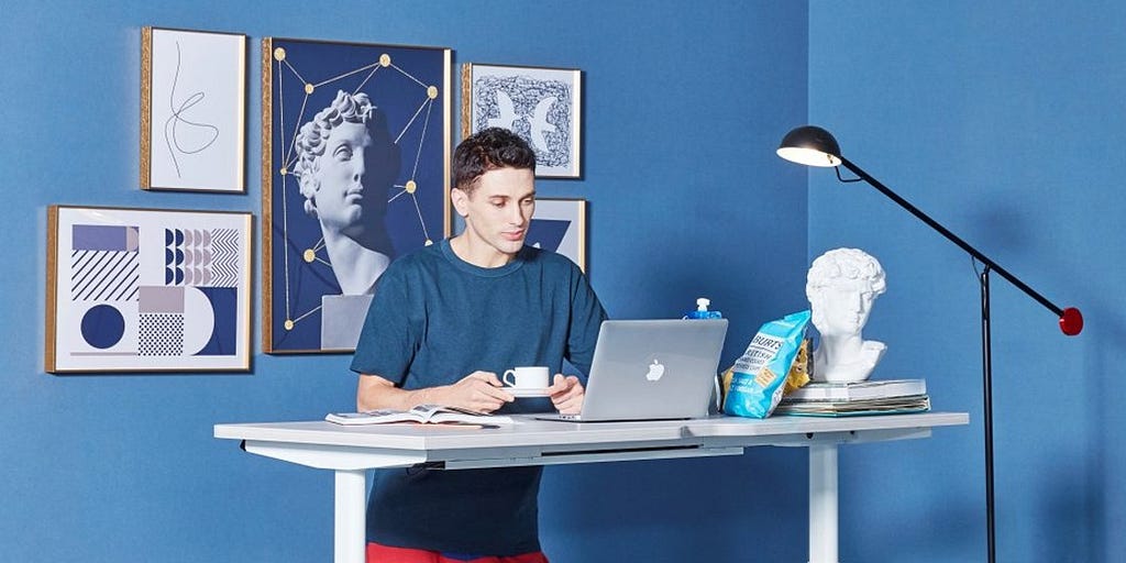 A Gen Z worker who is working from his home office. He is in a room with blue walls and picture frames hung on the wall behind him. He is standing as a standing desk looking at his laptop and drinking his cup of hot beverage. He is wearing casual clothes of a blue t-shirt and red sweat pants..