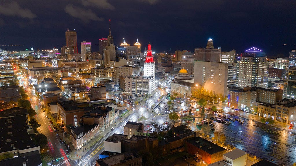 Aerial nighttime view of downtown Buffalo, New York, showcasing the city’s bright lights and urban landscape. The image captures the busy streets with light trails from moving vehicles, illuminated buildings, and a clear view of the city’s architecture, including prominent buildings lit in red and a distinctive dome-shaped structure. The urban grid layout is visible, and the atmosphere is vibrant with a mix of residential and commercial areas.