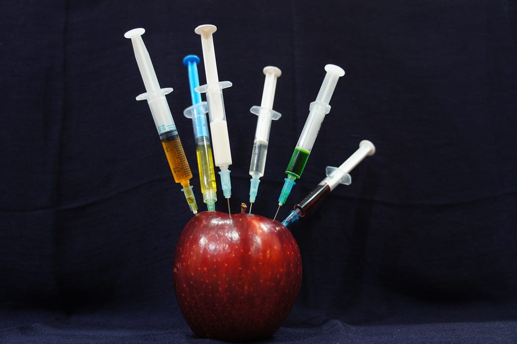Photo of an apple with 6 different coloured syringes sticking into it from different angles