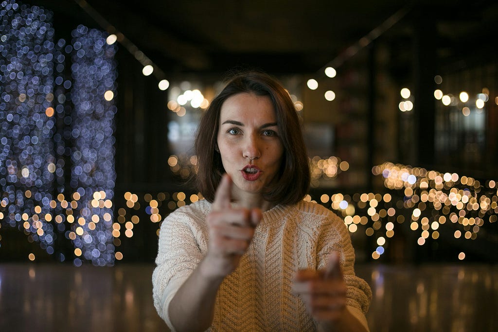 Woman with finger upraised speaking direct to camera. In the background, a city at night.