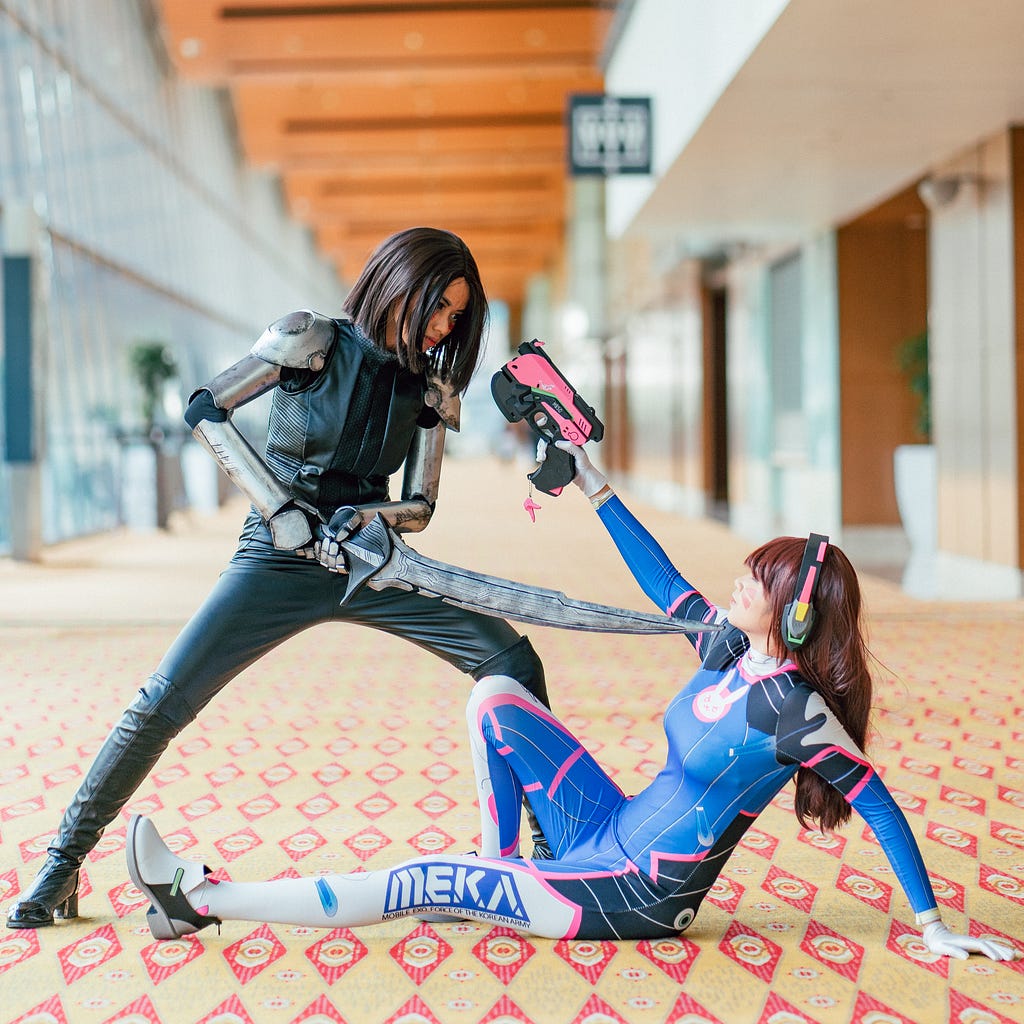 Two women wearing cosplay fighting — one stood up with a sword pointed at the face of the other, who is sat on the ground holding a gun to the first woman’s head