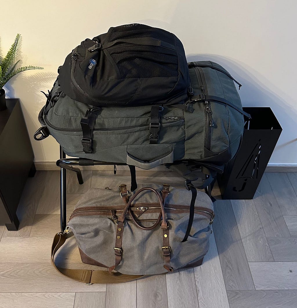 A light grey holdall with brown leather steps and a large dark grey travel backpack with smaller black detachable backpack attached.