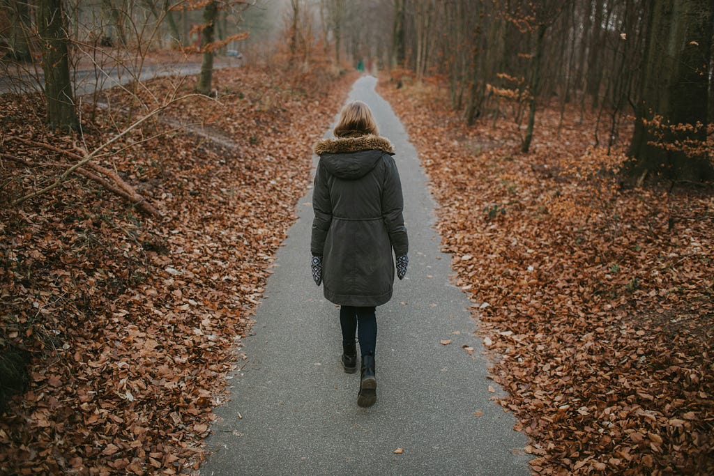 A woman in winter coat walks alone down a narrow path in the woods, seen from behind.