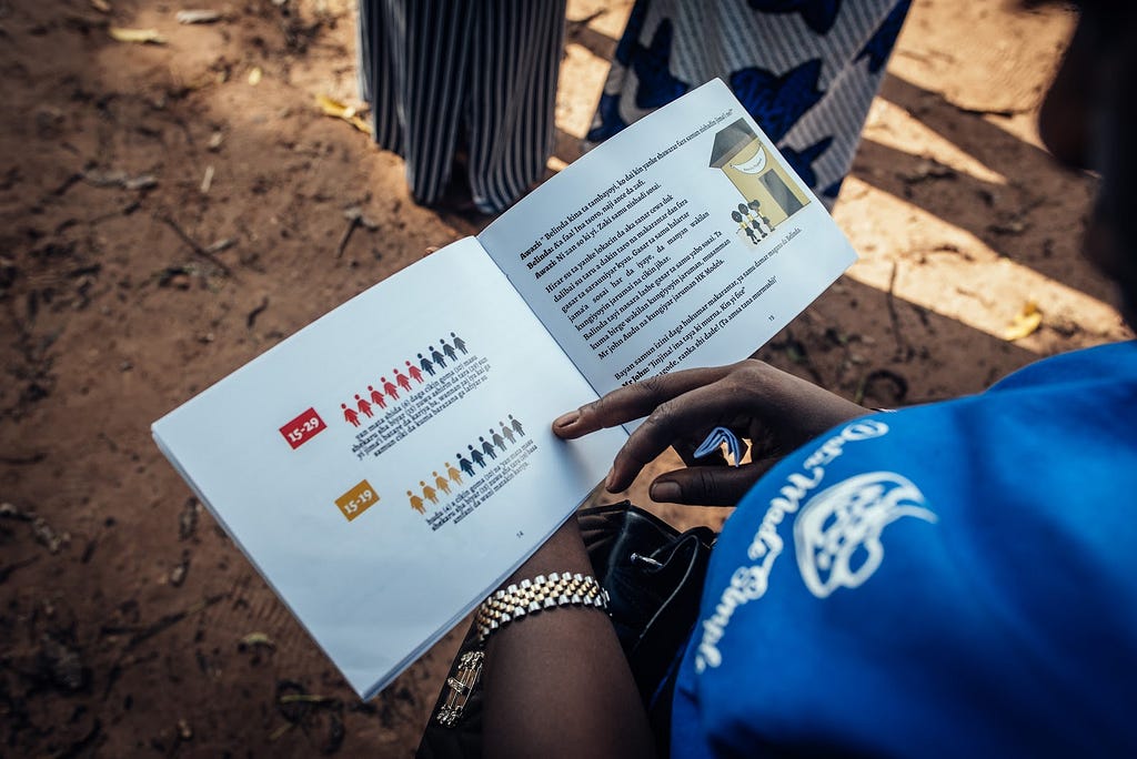 The camera is looking over the shoulder of a community member, at the Data Made Simple storybook that they are holding. The pages display simple data visualizations and graphics, with text in a local Nigerian language.