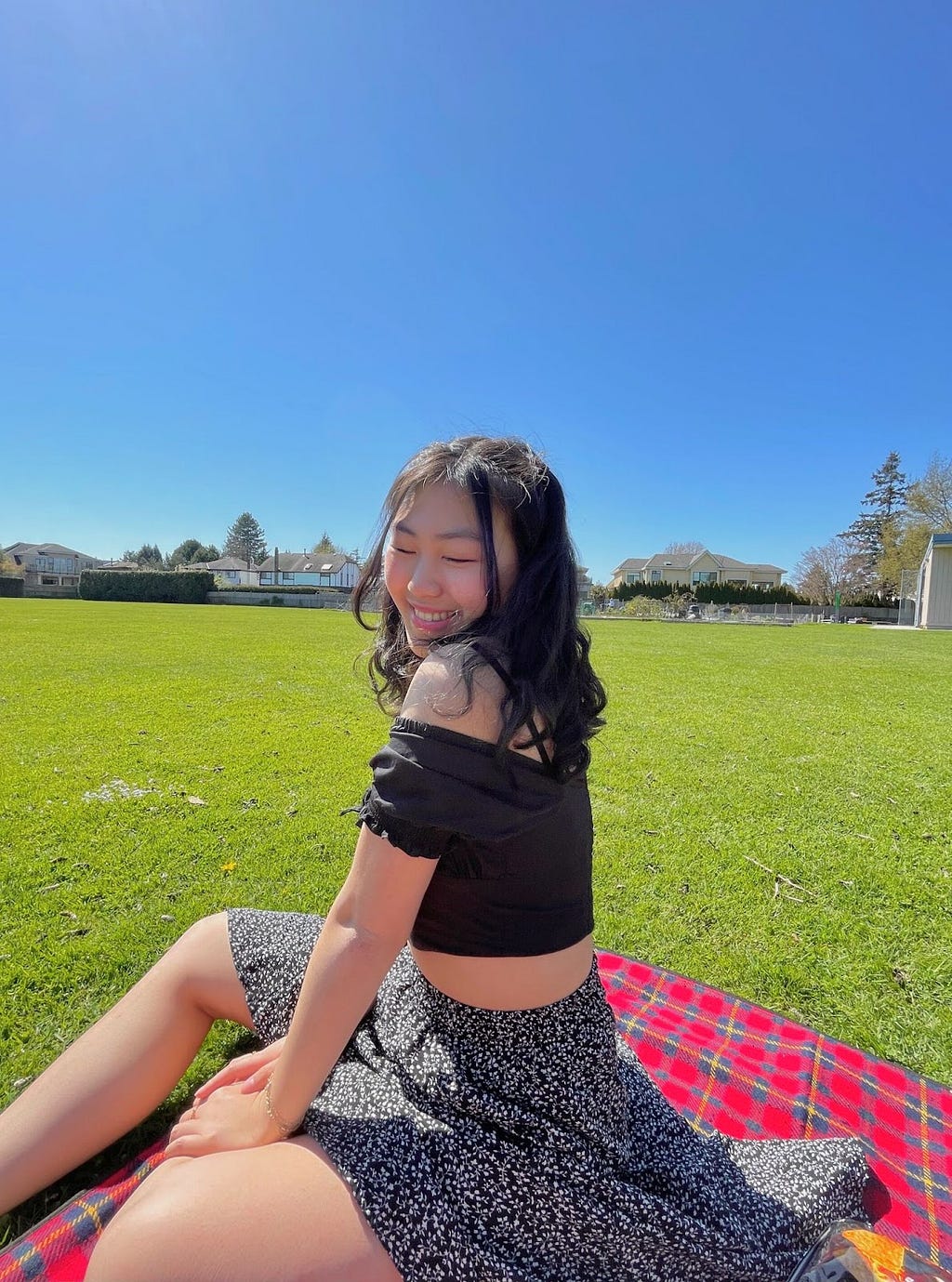 Photograph of smiling Chinese-American woman with black hair. She is sitting on a grassy field, wearing a black top and a long black floral skirt.