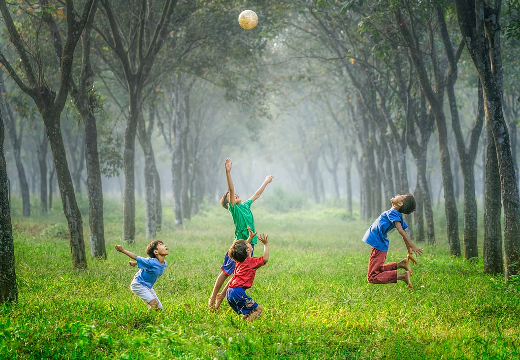 Four boys in colorful red, green, dark blue and pale blue T-shirts and shorts trying to catch a white ball high over their heads , in a grassy field with a line of tall green trees on both sides of the field that fades into a foggy background.