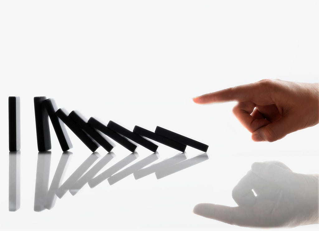 A picture of nine black dominoes lined up vertically and a finger on a hand knocking over the first domino, resulting in the rest of the dominoes falling in sequence.