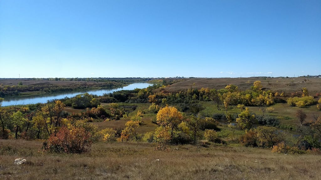 Foto de paisagem com gramado e árvores, parte da vegetação está amarela e avermelhada de outono, há um rio na parte esquerda e o céu está limpo e azul