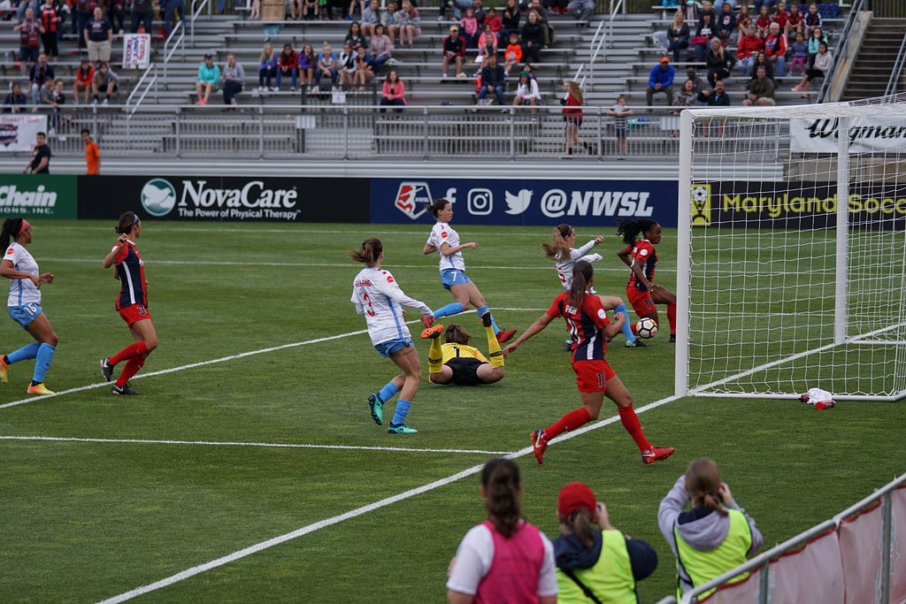A female soccer–player in red scoring a goal right in the penalty area against a team dressed in white.