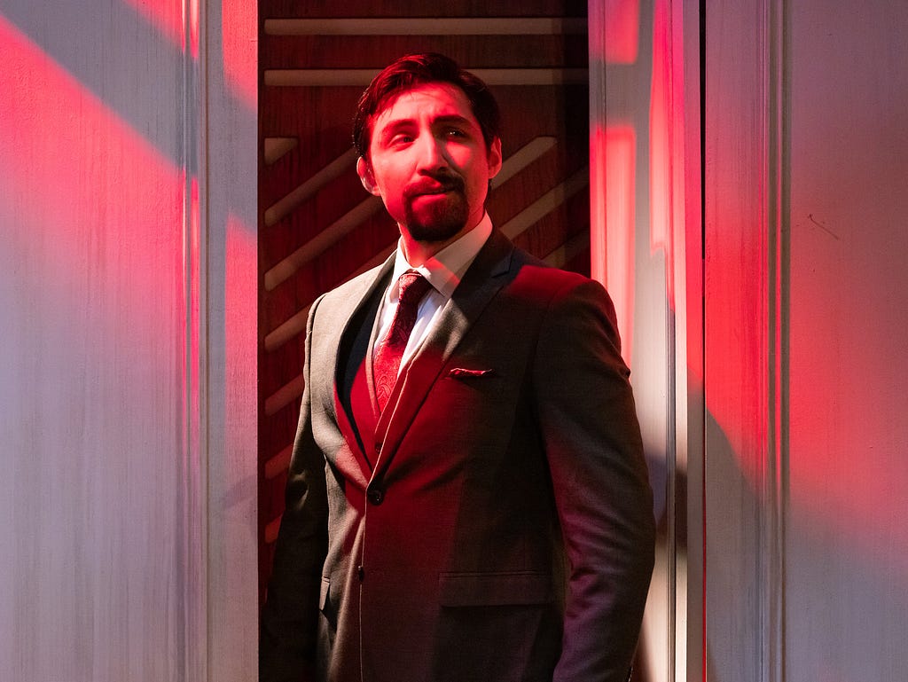 A man with tan skin, dark brown hair, and a dark brown goatee stands in a dark suit, bathed in red light.