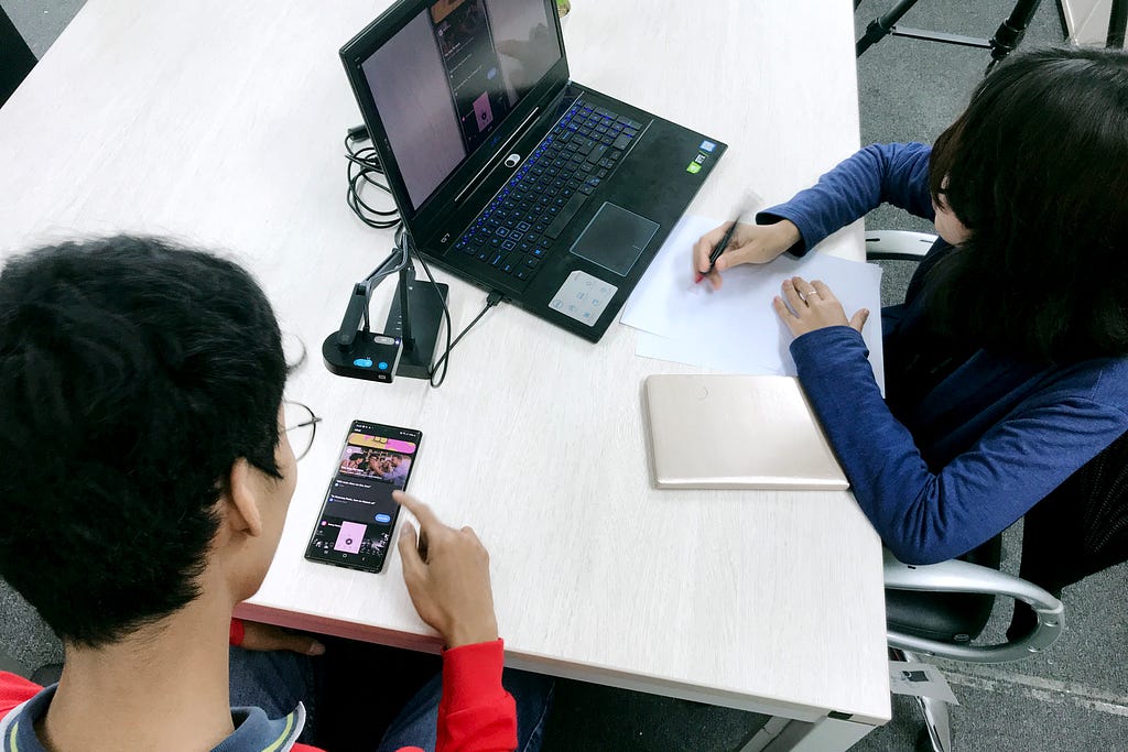 A young man looks at his phone at a desk next to a woman writing on a piece of paper in front of her laptop.