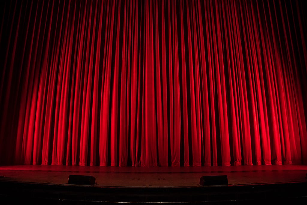 first person view of a stage. the stage has a wood floor and big bright red curtains.