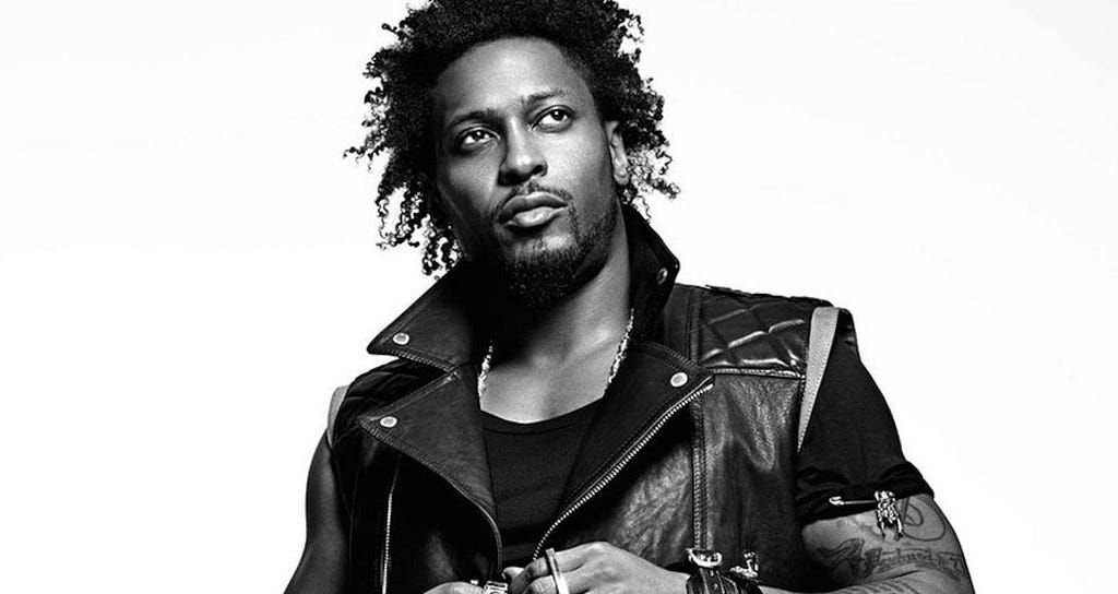 A black-and-white photo of Black musician D’Angelo. In the picture, he is wearing a black leather vest on top of a black T-shirt. His hair is out in a curly afro.