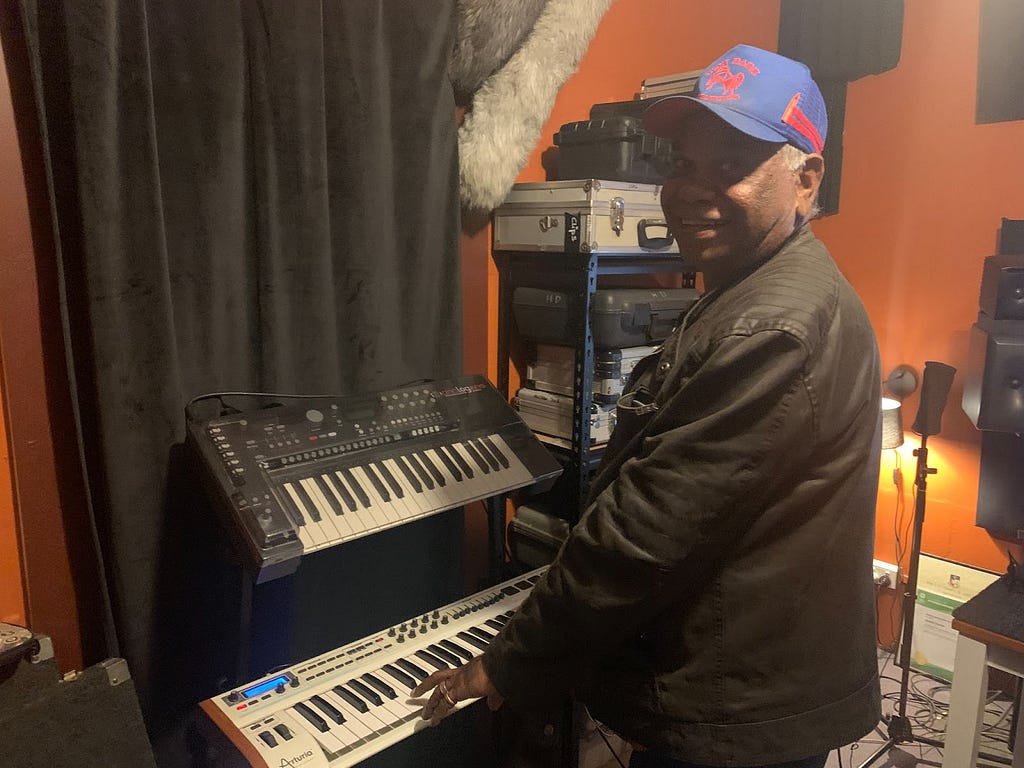 A photograph of a man smiling at the camera while playing a keyboard in a room with musical equipment.