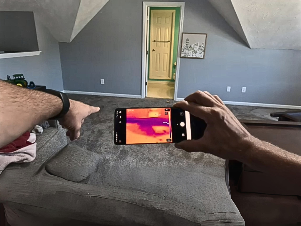 Professional conducting a thermal imaging inspection using a smartphone with a thermal camera app, pointing at a suspected water damage area on a grey carpet, indicating potential for water removal or mold remediation services.