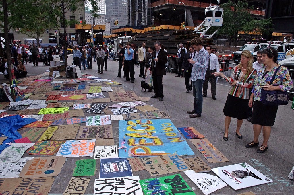 Photos of onlookers wearing business wear pointing at an array of colorful signs that say things like “GREED KILLS” and “PEACE NOW” from Zuccotti Park, September 28 2011.