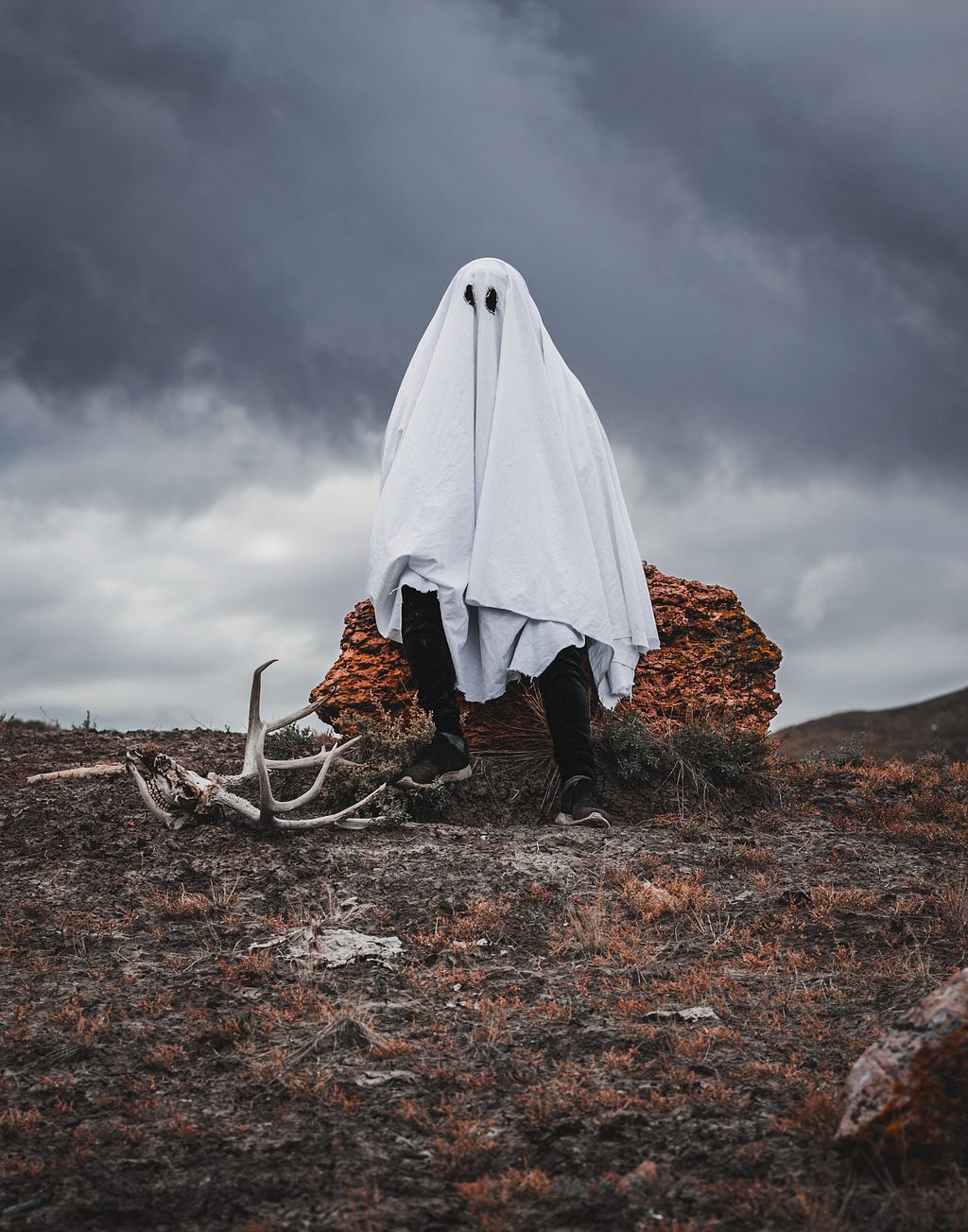 Uma pessoa fantasiada de fantasma, sentada em uma pedra laranja em um tipo de deserto. O céu está cheio de nuvens escuras