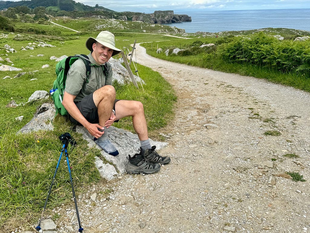 The author sitting on a rock, with one boot off, next to a gravel road besides the sea.