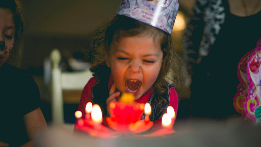 Picture of a young girl seated at a table in a party hat with her mouth wide open, either screaming or gathering breath to blow out 6 red candles on her birthday cake.