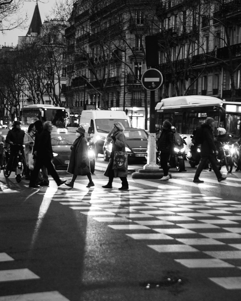 A black and white image of a busy street with people crossing it at a Zebra Zrossing and many vehicles waiting on the other side.