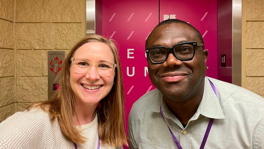 Natasha, a white woman with red shoulder length hair and clear glasses, and Zuby, a Black man with short hair and black glasses, smile in front of the elevator heading up to the Remarkable Tech Summit