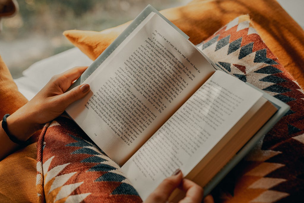 Cozy photo of a person’s hands holding a book while resting on a warm-toned pillow with geometric print.
