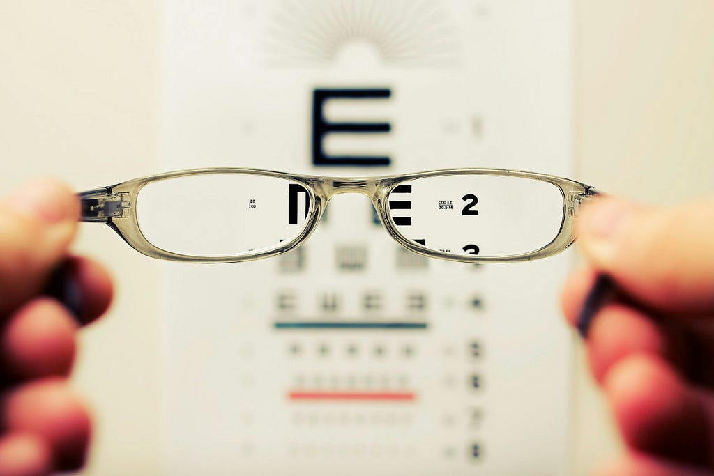 Eye exam, a pair of glasses being held in front of various sizes of E and being blurry except for in the glasses lenses