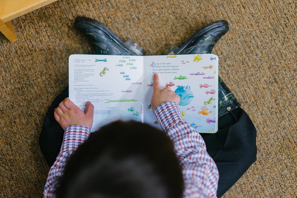 Uma criança, sentada em um tapete marrom claro, lendo um livro infantil