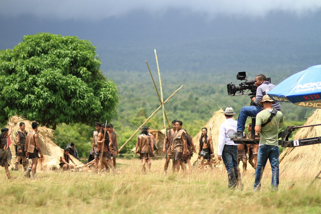 A camera crew dressed in modern clothing and using modern equipment is filming what appears to be a tribal scene or primitive scene. The full picture of the scene gives us perspective we might not have from an angel where we could only see one of the two groups.