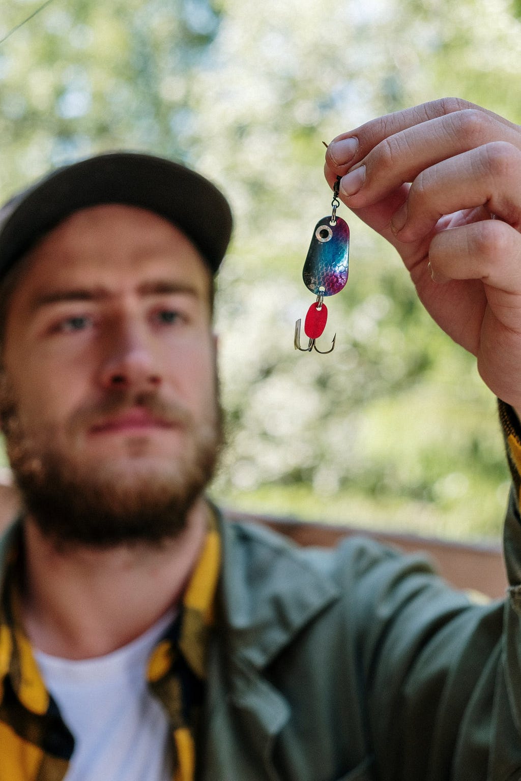 A shimmery blue and purple metal fishing lure with several hooks hanging from it is being held up close to the camera by a somewhat out of focus  white bearded male wearing a green jacket with a black and yellow plaid shirt underneath.