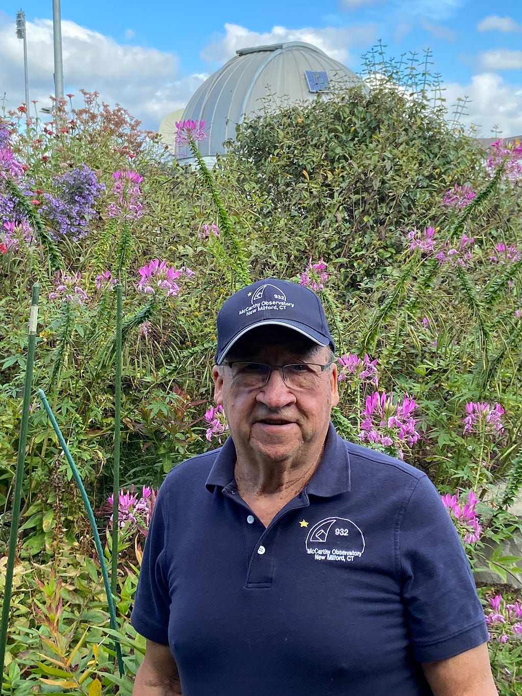 Bob Lambert, an older gentleman, wears a blue baseball cap and blue polo shirt, and stands in front of native flowers at the observatory.