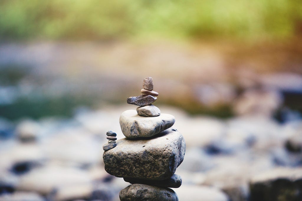 Stack of greyish rocks, one big rock has moss on it, larger rocks in a central stack, and some smaller ones on top, a few little ones stacked off to one side, a long tiny one in the stack of smaller ones on top. With blurred background of a riverbed and some bushes.