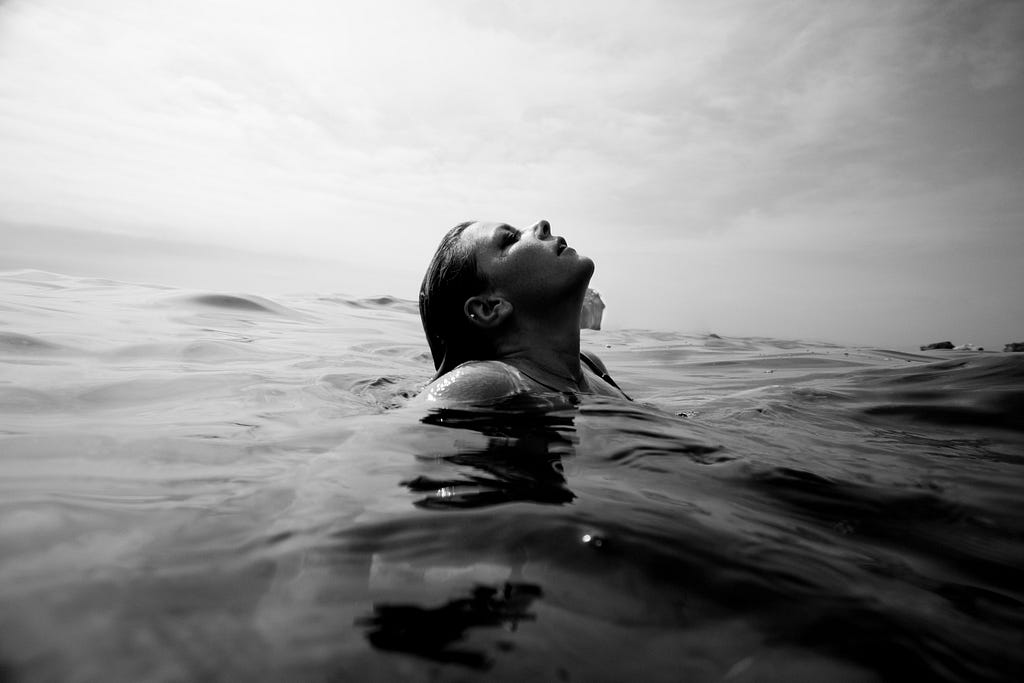 Foto em preto e branco de uma mulher saindo do fundo do mar para respirar. Sua postura sugere que está aliviada por ter conseguido recuperar o folego.