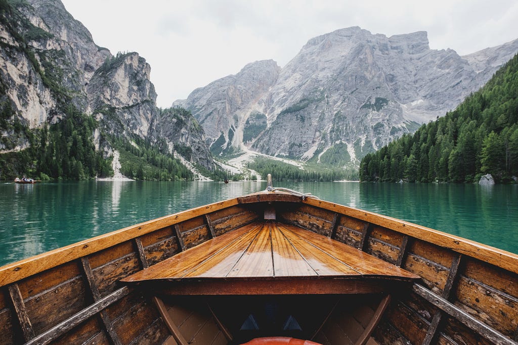 A view of mountains, forest, and a lake, from the position of someone seated in a wooden boat on the lake.