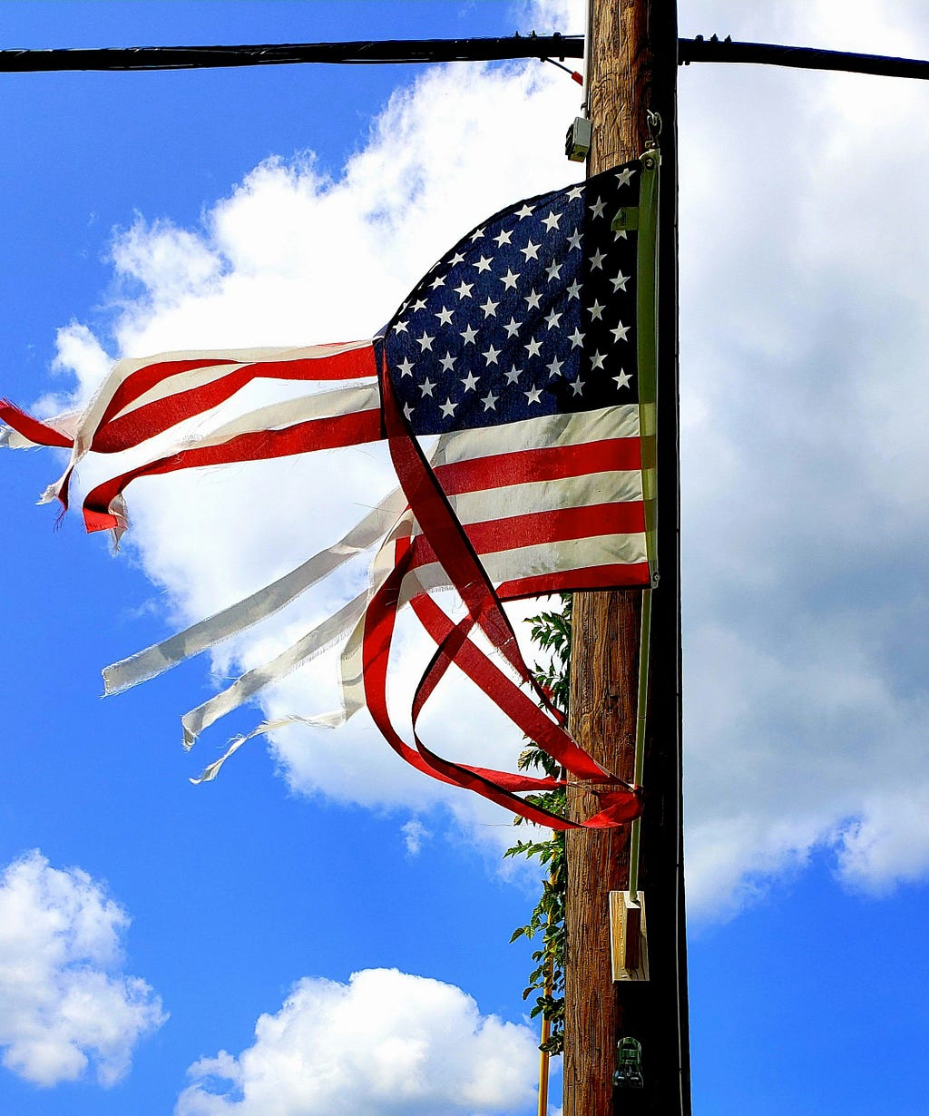 Centered is a shredded American flag mounted on a power pole-the pole and perpendicular power lines are suggestive of a christian cross.