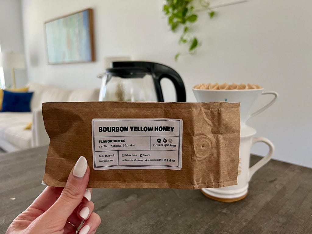 A hand holds up a bag of coffee labeled “Bourbon Yellow Honey.” In the background is a coffee mug, coffee pot, and furniture.