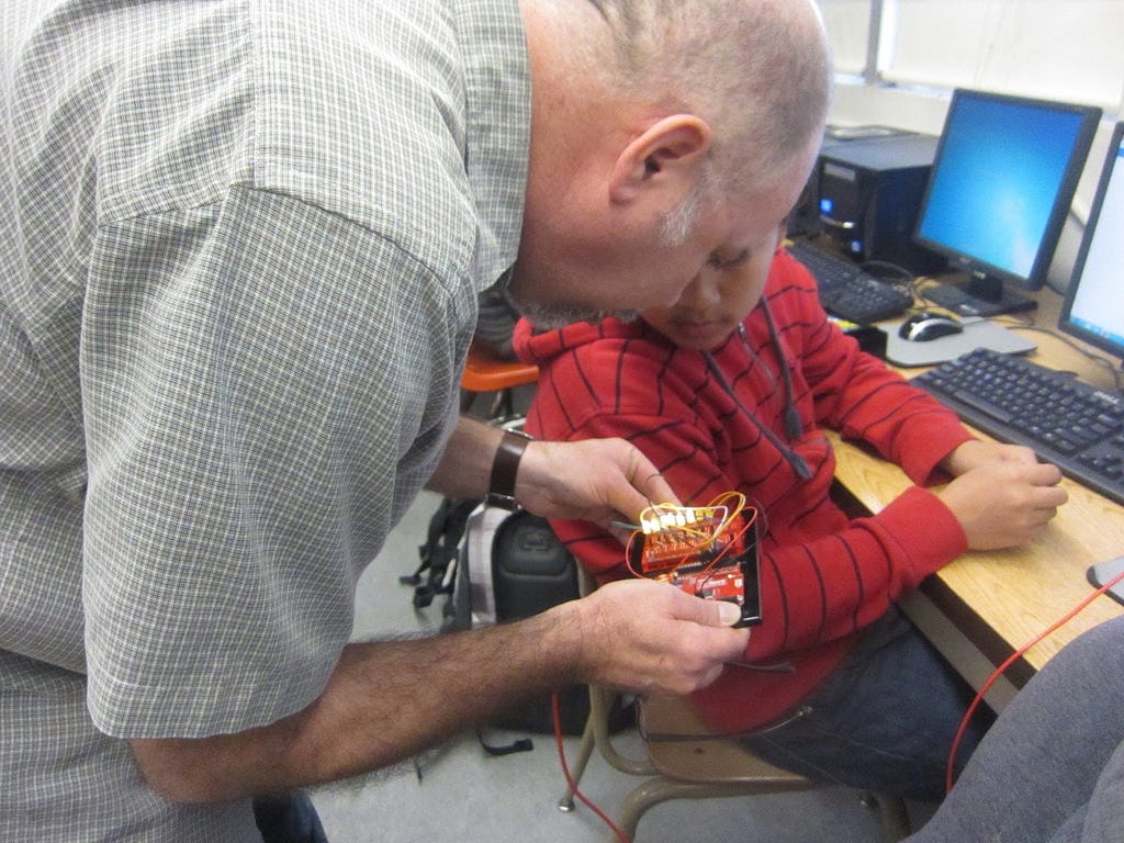 A man is standing and leaning down to look at the electrical project in his hands. A seated student is also looking at it.