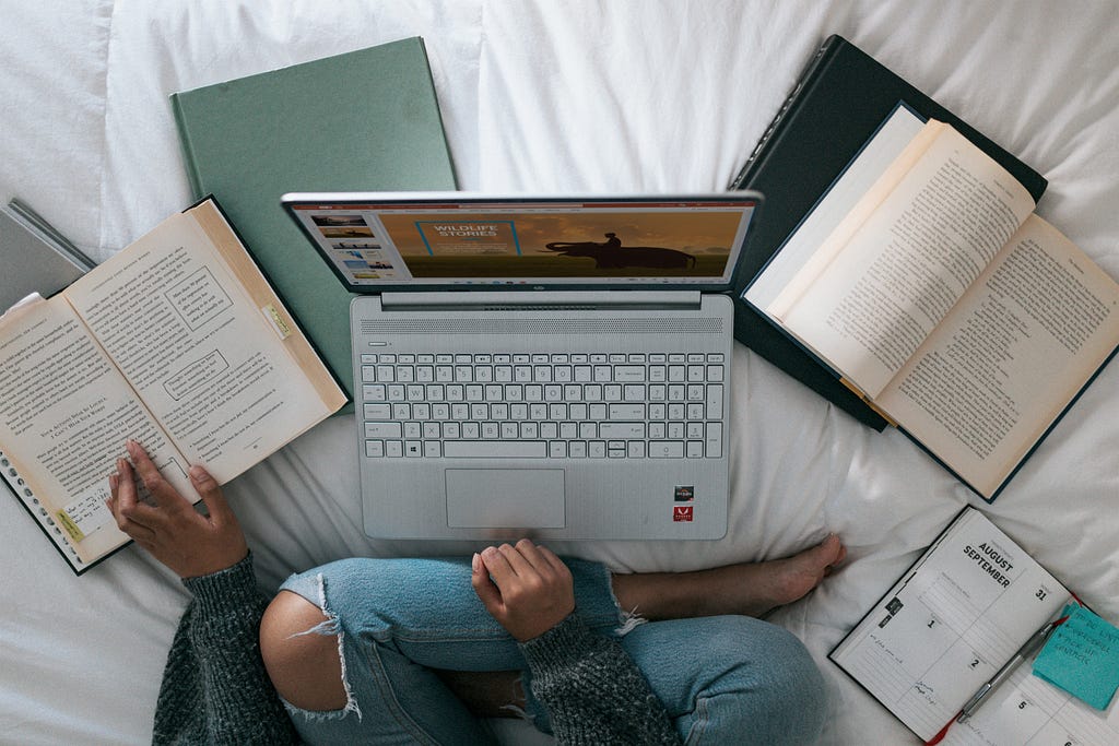 A person sitting on a bed with laptop, books, and notes. They’re studying.