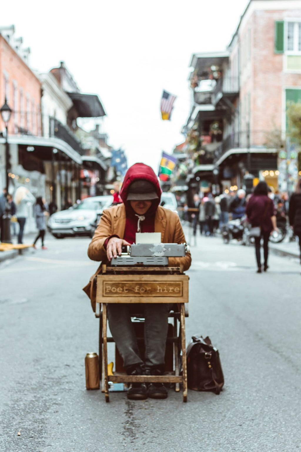 Photo of a person sitting in the middle of the street at a small wooden desk that says “Poet for hire” on the front. The person is using a typewriter that is on top of the desk.