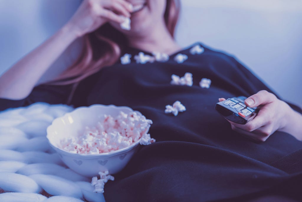 A woman watches tv, laughing and eating pop corn