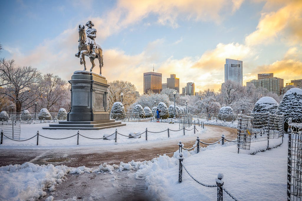 A scene from the Boston Public Garden of a statute of a man on a horse covered in snow with the city in the background.