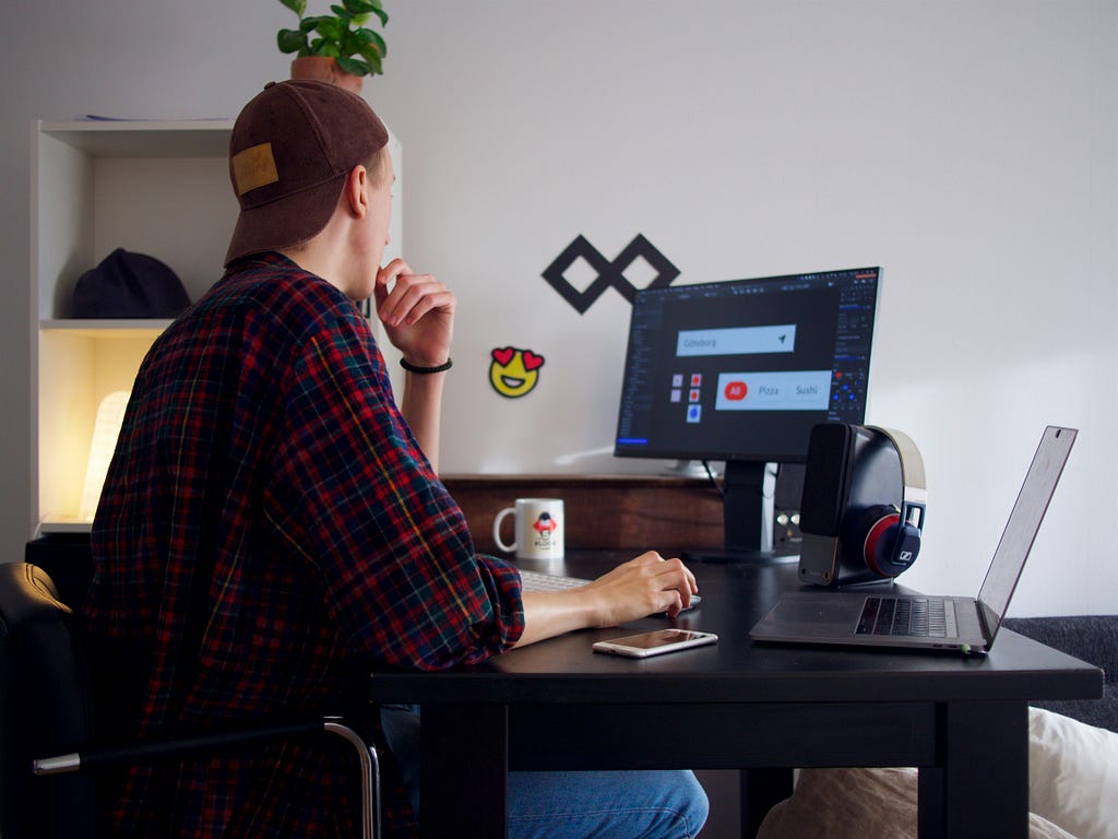 A young freelancer wearing a hat looking at his desktop screen with a laptop on the side