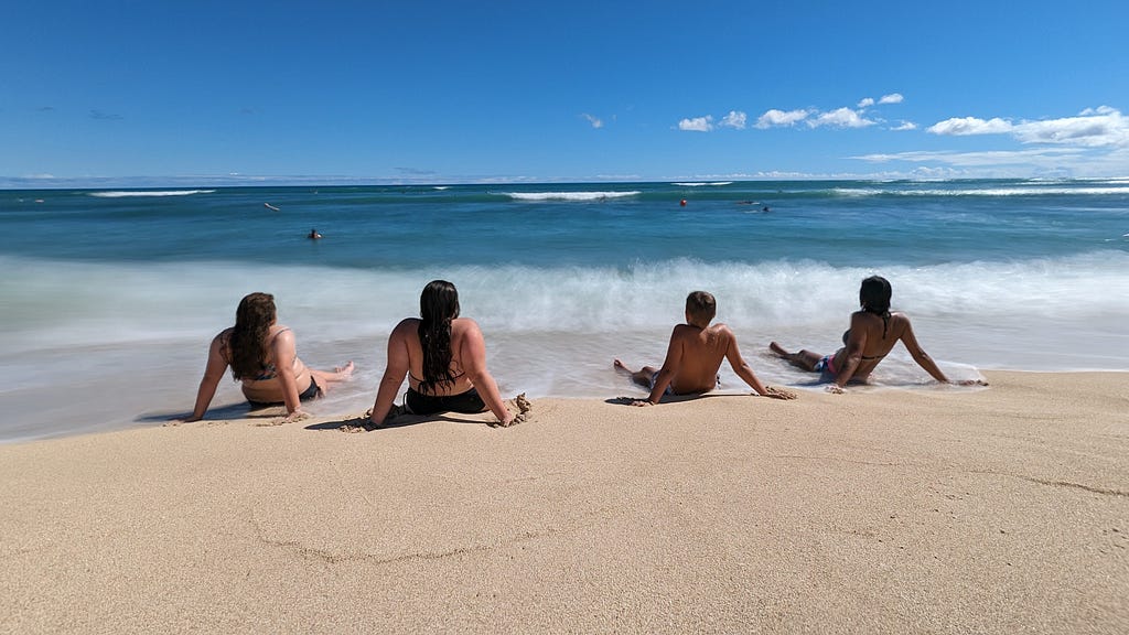 Four people are sitting on the sandy shore with their backs to the camera, facing the ocean. The water is a vibrant blue with gentle waves under a clear, sunny sky. As they ponder &quot;How Many More Summers?&quot; other individuals can be seen swimming and enjoying the water in the distance.