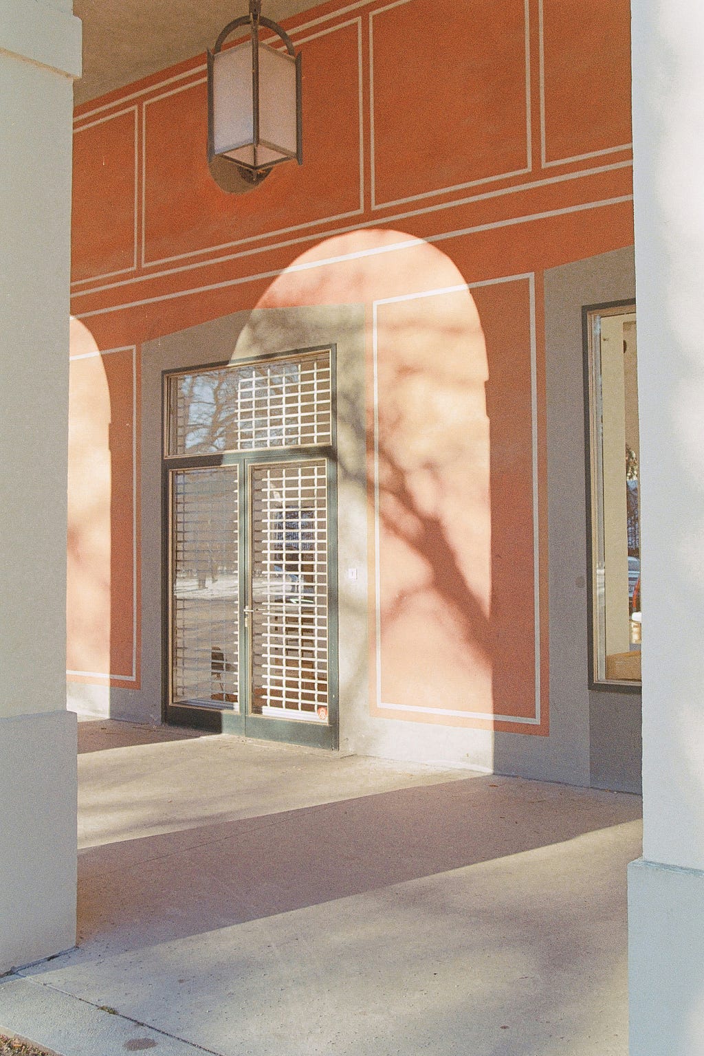A section of a building’s exterior with warm orange walls and paler orange geometric trim, featuring a hanging lantern, grid-pane windows, and door, with the shadow of a tree cast upon the wall.