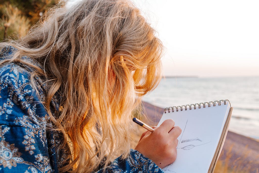 A profile view of a woman with a notebook and pen, sitting on a beach in sunlight