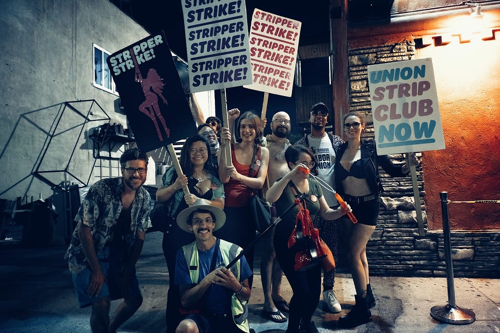 A group of video game workers and allies pose for a photo in front of the Star Garden Dive Bar. Workers hold colorful signs reading “STRIPPER STRIKE” and “UNION STRIP CLUB NOW.” Amazon Labor Union president Chris Smalls is also present
