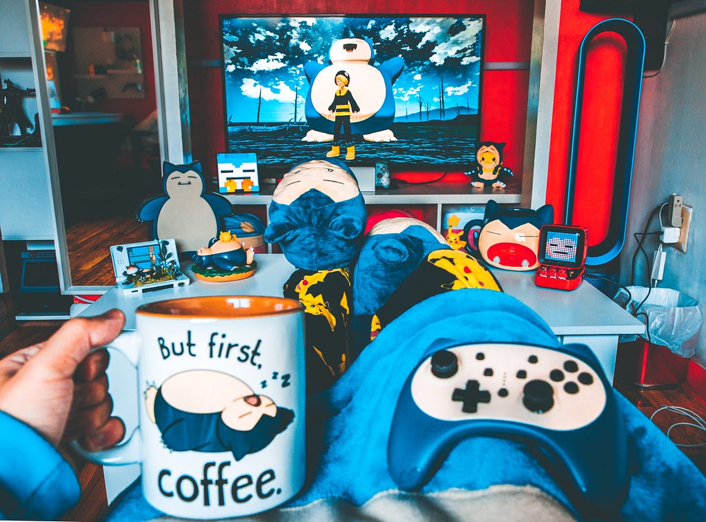 A room from a person Point of View with coffee mug that says — But first Coffee, then a gaming console, TV, and stuff toys of Snorlax in the background